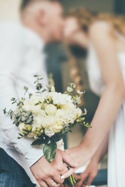 Yeni evlilerin elinde bir düğün buketinin yakın çekimi. Dikey fotoğraf. Arka planda, gelin ve damat öpüşüyorlar. Yüksek kalite fotoğraf