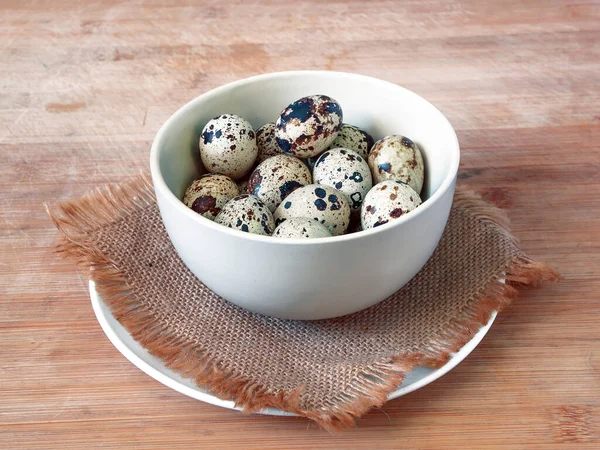 The quail eggs in the ceramic bowl on the jute rug and ceramic plate on the wooden table. Closeup.