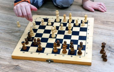 Sıradan giyinmiş beyaz bir çocuk ahşap zeminde oturuyor ve eski bir satranç tahtasıyla satranç oynuyor..