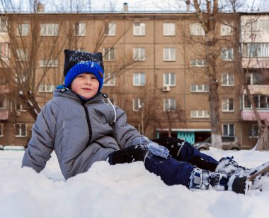 Yeşil ceketli ve mavi örgü şapkalı gülümseyen beyaz çocuk kışın açık havada karda oturuyor..