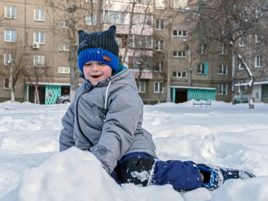 Yeşil ceketli ve mavi örgü şapkalı gülümseyen beyaz çocuk kışın açık havada karda oturuyor..