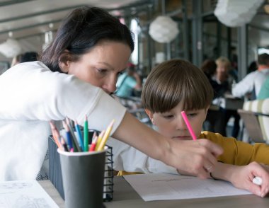 Annesiyle birlikte bir sokak kafesindeki masada oturan bir çocuk kalemle resim çiziyor..