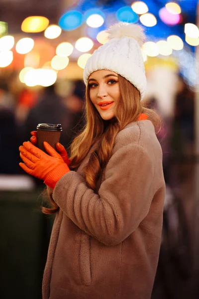 Vacker flicka på bakgrunden av ljus med kaffe, på gatan, semester, nytt år. en promenad genom kvällsstaden — Stockfoto