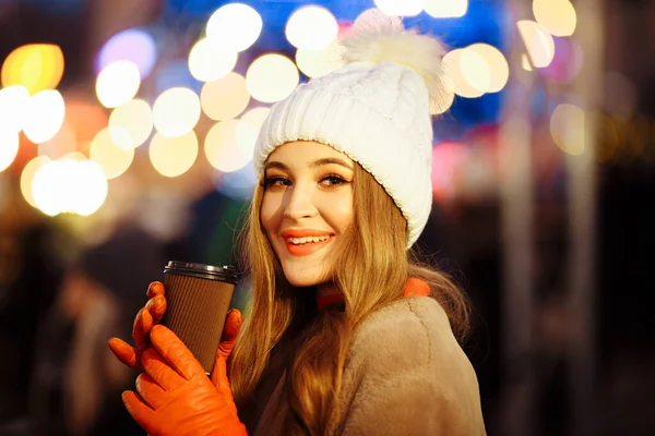 Gadis cantik di latar belakang lampu dengan kopi, di jalan, liburan, tahun baru. berjalan-jalan melalui kota malam — Stok Foto