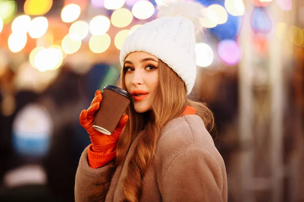 Vacker flicka i en vit hatt och beige päls, på kvällen, på vintern, dricker en drink från ett glas mot bakgrund av kvällsljus. — Stockfoto