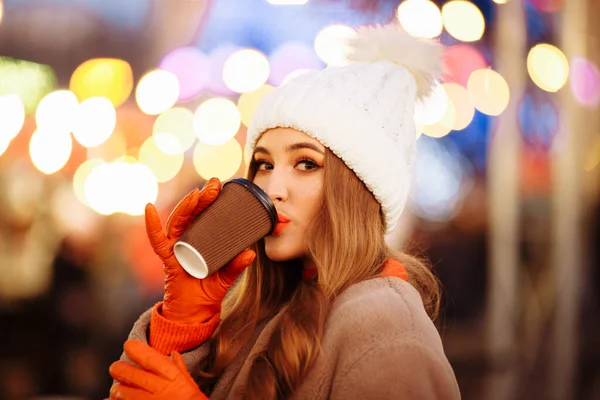 白い帽子とベージュの毛皮のコートに身を包んだ美少女が冬の夜には夜の光を背景にガラスから飲み物を飲み. — ストック写真