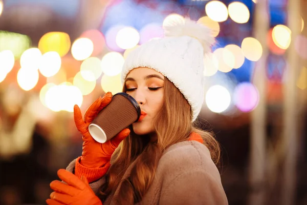 Gadis cantik dengan topi putih dan mantel bulu beige, di malam hari, di musim dingin, minum dari gelas dengan latar belakang lampu malam. — Stok Foto