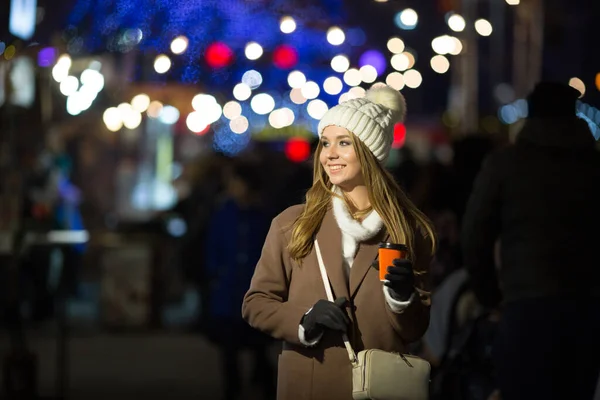 Belle fille, le soir, sur fond de lumières avec une boisson dans un verre orange, un chapeau blanc et un manteau beige avec un sac. — Photo