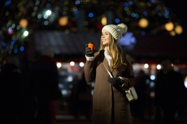 Güzel kız, akşamları, ışıkların arka planında, turuncu bardakta bir içkiyle, beyaz şapkalı ve çantalı bej ceketli..