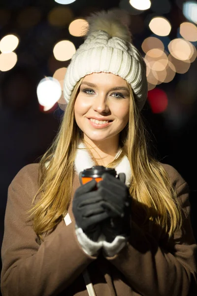 美しい少女は、夕方には、オレンジのガラス、白い帽子とベージュのコートでドリンクとライトの背景に。カメラで笑って — ストック写真