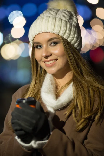 Vacker flicka, på kvällen, mot bakgrund av ljus med en drink i ett orange glas, en vit hatt och en beige rock med en väska. — Stockfoto