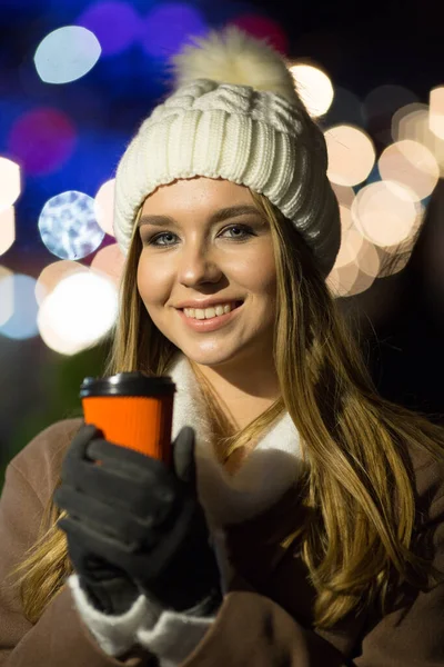 Menina bonita, à noite, no fundo das luzes com uma bebida em um copo de laranja, um chapéu branco e um casaco bege. sorrindo para a câmera — Fotografia de Stock