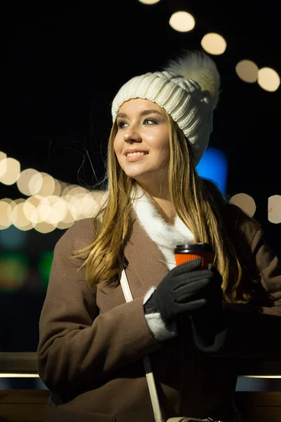 Menina bonita, à noite, no fundo das luzes com uma bebida em um copo de laranja, um chapéu branco e um casaco bege com uma bolsa. — Fotografia de Stock