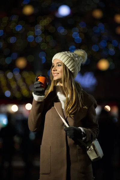Belle fille, le soir, sur fond de lumières avec une boisson dans un verre orange, un chapeau blanc et un manteau beige avec un sac. — Photo