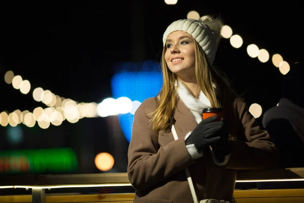 Uma menina loira, à noite, no fundo das luzes da feira com uma bebida em um copo de laranja, um chapéu branco e um casaco bege caminha na rua. — Fotografia de Stock