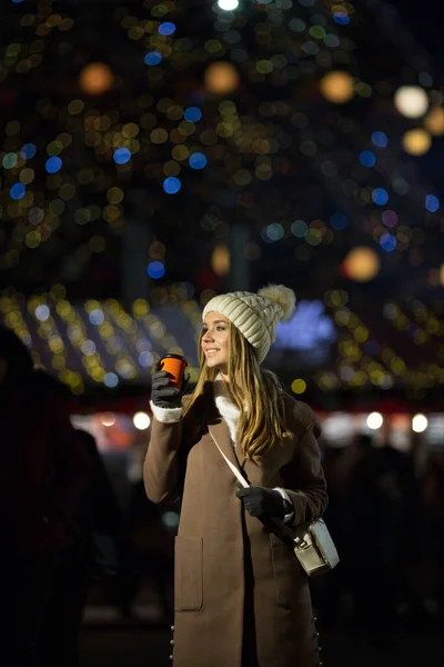 Una chica rubia, por la noche, sobre el fondo de las luces de la feria con una bebida en un vaso naranja, un sombrero blanco y un abrigo beige camina por la calle. —  Fotos de Stock