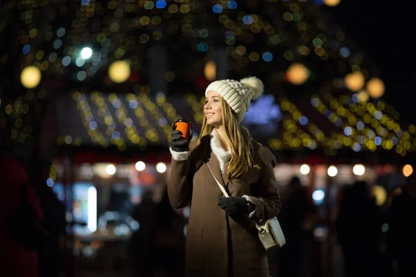 Une fille blonde, dans la soirée, sur le fond des lumières de la foire avec un verre orange, un chapeau blanc et un manteau beige marche dans la rue. — Photo