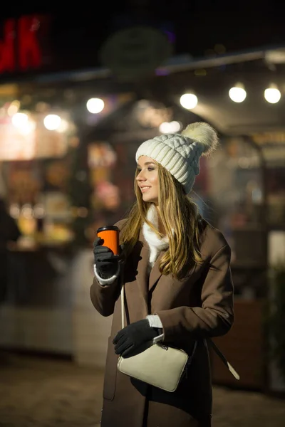Uma menina loira, à noite, no fundo das luzes da feira com uma bebida em um copo de laranja, um chapéu branco e um casaco bege caminha na rua. — Fotografia de Stock
