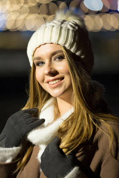 Belle fille, le soir, sur fond de lumières, un chapeau blanc et un manteau beige. sourire à la caméra — Photo