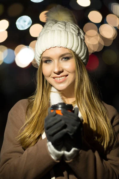 Uma menina loira, à noite, no fundo das luzes da feira com uma bebida em um copo de laranja, um chapéu branco e um casaco bege caminha na rua. — Fotografia de Stock