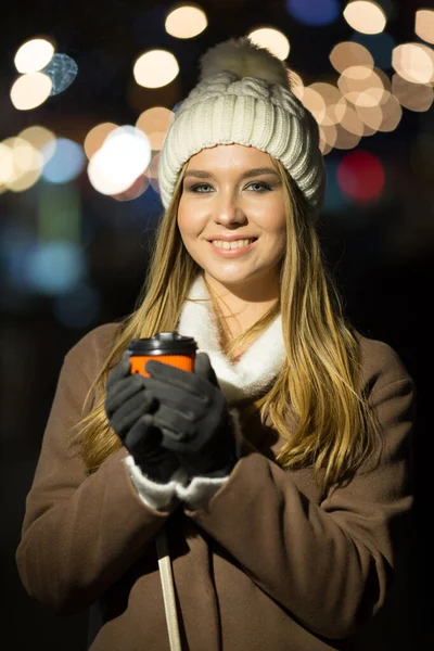 Uma menina loira, à noite, no fundo das luzes da feira com uma bebida em um copo de laranja, um chapéu branco e um casaco bege caminha na rua. — Fotografia de Stock