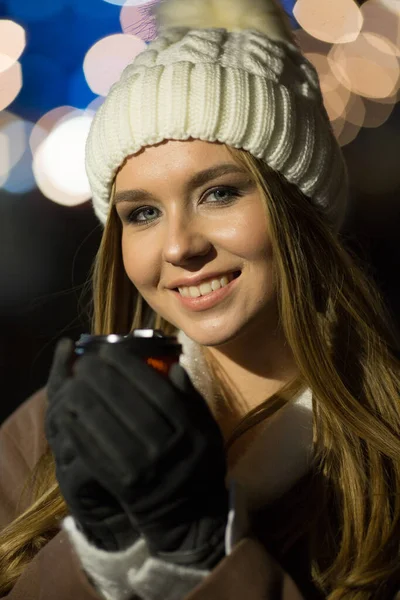 夜になるとブロンドの女の子がオレンジのガラスのドリンクと白い帽子とベージュのコートを背景に街を歩き. — ストック写真