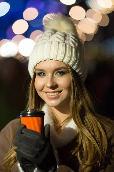 En blond flicka, på kvällen, mot bakgrund av ljusen på mässan med en drink i ett orange glas, en vit hatt och en beige rock promenader på gatan. — Stockfoto