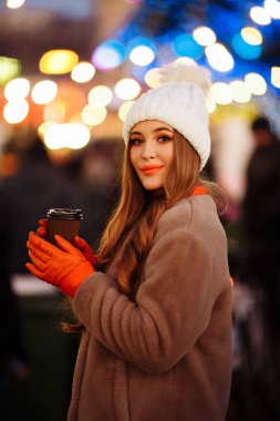 Işıkların arka planında güzel bir kız, kahveyle, sokakta, tatilde, yeni yılda. Akşam kentinde bir yürüyüş.