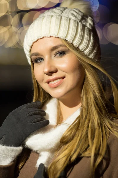 Menina bonita, à noite, no fundo das luzes, um chapéu branco e um casaco bege. sorrindo para a câmera — Fotografia de Stock