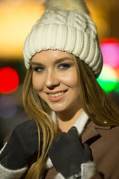 Hermosa chica, por la noche, sobre el fondo de luces, un sombrero blanco y un abrigo beige. sonriendo a la cámara —  Fotos de Stock