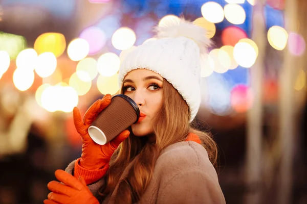Gadis cantik di latar belakang lampu dengan kopi, di jalan, liburan, tahun baru. berjalan-jalan melalui kota malam — Stok Foto