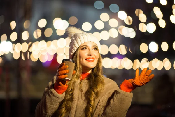 Vacker flicka på bakgrunden av ljus med kaffe, på gatan, semester, nytt år. en promenad genom kvällsstaden — Stockfoto