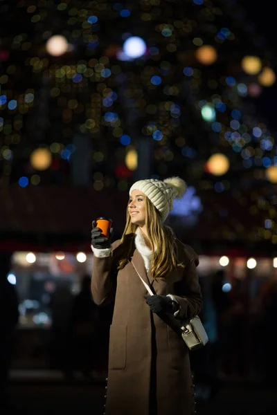美しい少女は 夕方には オレンジのガラス 白い帽子とベージュのコートでドリンクとライトの背景に カメラとクローズアップのポートレートで笑っています — ストック写真