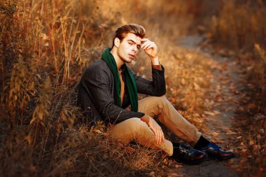 Gri ceketli bir adam, yeşil atkı, bej pantolon ve sonbaharda çimlerin üzerinde oturan bir süveter. Poz veriyor. Düşüncelilik ve üzüntü.