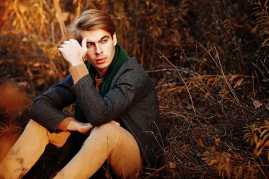 Gri ceketli bir adam, yeşil atkı, bej pantolon ve sonbaharda çimlerin üzerinde oturan bir süveter. Poz veriyor. Düşüncelilik ve üzüntü.