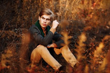 Gri ceketli bir adam, yeşil atkı, bej pantolon ve sonbaharda çimlerin üzerinde oturan bir süveter. Poz veriyor. Düşüncelilik ve üzüntü.