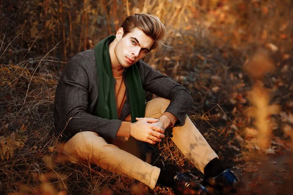 Muž, model v šedém saku, zelený šátek a béžové kalhoty a svetr sedí na podzim na trávě mezi podzimními stromy. pózování. přemýšlivost a smutek. — Stock fotografie