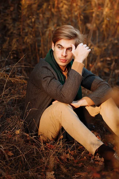 a man, a model in a gray jacket, a green scarf and beige pants and a sweater sits on the grass in autumn, among the autumn trees. posing. thoughtfulness and sadness.