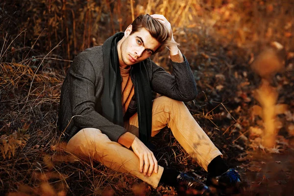Мужчина, модель в серой куртке, зеленый шарф и бежевые брюки и свитер осенью сидят на траве, среди осенних деревьев. позирует. вдумчивость и печаль. Лицензионные Стоковые Изображения