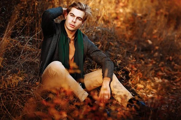 A man, a model in a gray jacket, a green scarf and beige pants and a sweater sits on the grass in autumn, among the autumn trees. posing. thoughtfulness and sadness. Stock Picture