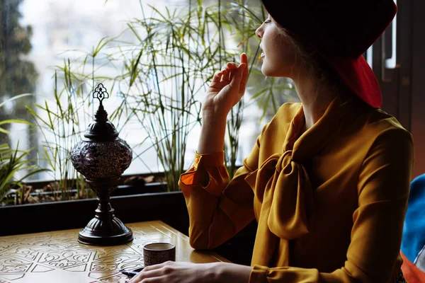 Chica en un café con una taza de café y un hat.portrait de la joven sensual con sombrero floppy y blusa con lazo. Hermosa mujer morena en el café sosteniendo la taza de café — Foto de Stock