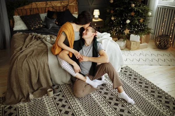 Счастливая влюбленная пара по утрам на кровати возле елки, объятия, рождественское утро. домашняя одежда и свитера. утешение и тепло сердец Лицензионные Стоковые Фото