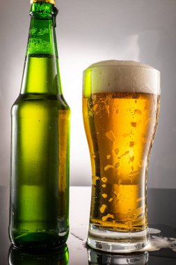 Bira bardağı, köpük ve yanında bira olan yeşil şeffaf şişe.