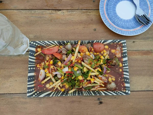 Tahta bir masada ekşi mangolu karides salatası. Sağ tarafta çay tabağı, sol tarafta su bardağı var. Bunlar Tayland 'da sokak yemekleri..