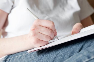 Kot pantolonlu ve beyaz tişörtlü bir kız not defterine kalemle yazıyor, yakın plan. Ev ya da uzaktan öğrenim kavramı, evden çalışmak, serbest çalışmak.