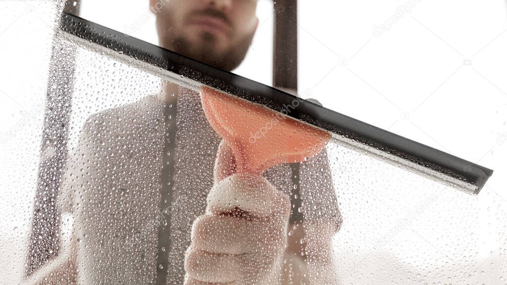 A male washes windows at home. House cleaning. Washing dirty window glass detergent for window washing. Cleaning a glass with a squeegee, close up.