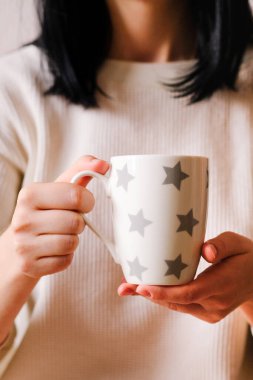 Elinde bir fincan kahve tutan rahat elbiseli kız. Hafif arka planda, sıcak içeceği olan kadın elleri.