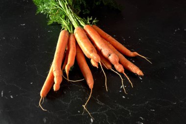 Tahta gibi siyah mermer üzerinde yeşil yaprakları olan organik çiğ havuç sürüsü, yukarıdan görünüyor.