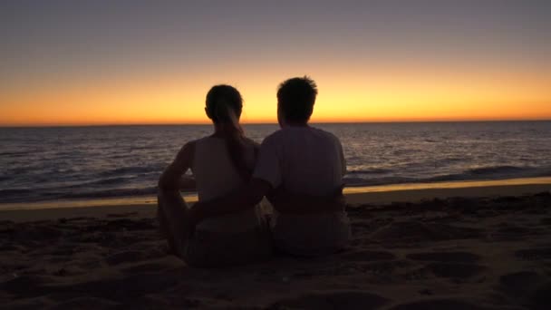 在日落后坐在海滩上 背景是平静的大海和橙色的天空 从后面看这对无名的年轻夫妇 — 图库视频影像