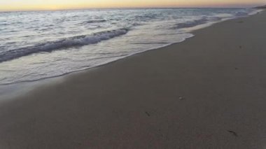 Gün batımından hemen sonra kumsal, kumları yıkayan küçük deniz dalgaları, arka planda turuncu gökyüzü.